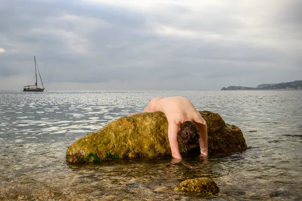 A conceptual fine art photograph by Arjan Spannenburg titled Siren, featuring a male figure slumped on a sea rock with a distant sailboat.