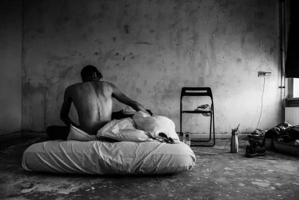A black and white fine art photograph titled Outcast showing a man sitting on a floor mattress in a desolate room.