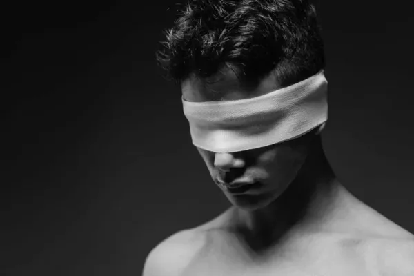 A minimalist black and white conceptual portrait of a man wearing a blindfold, emphasizing shadows and skin texture.