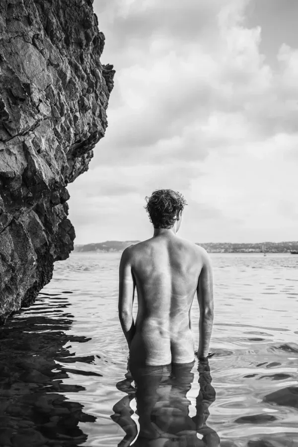 A serene black and white fine art photograph of a figure standing in water next to a cliff side, exploring themes of nature and vulnerability.