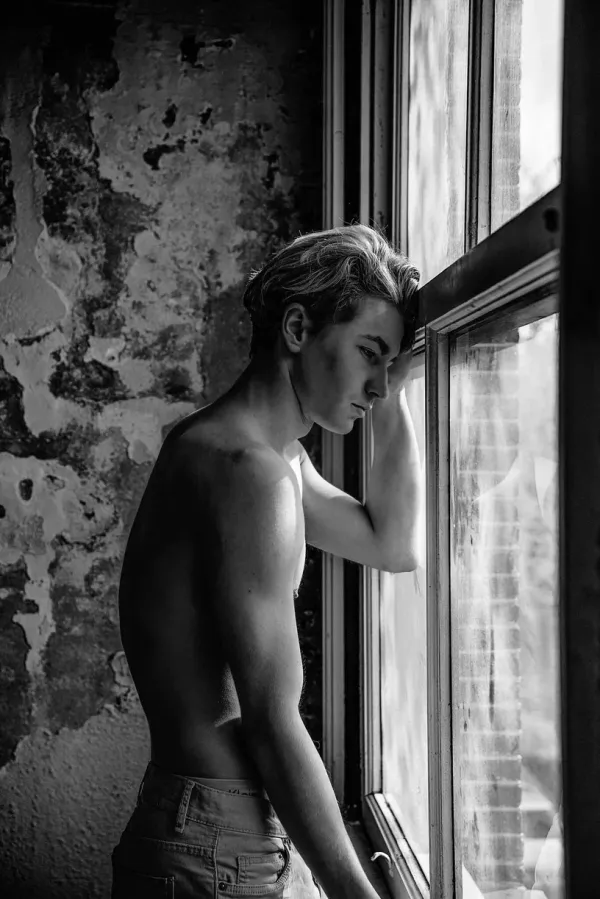 A black and white fine art portrait of a young man in profile, leaning his forehead against a window frame in a room with weathered, peeling walls. The lighting creates a high-contrast study of form and texture, capturing a moment of quiet introspection and vulnerability.