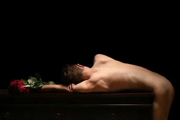 Fine art photograph by Arjan Spannenburg titled 'Kiss Goodbye,' featuring a vulnerable shirtless man reclining on a dark wooden cabinet beside a bouquet of red roses, symbolizing the numbing pain and exposure of lost love against a dramatic black backdrop.