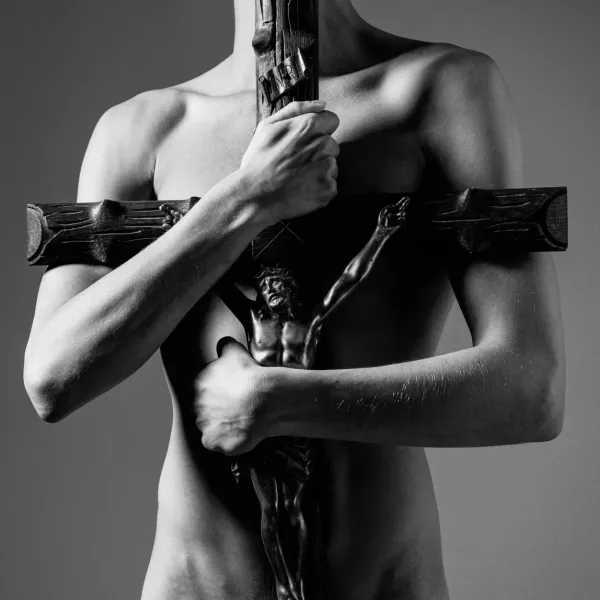 The artwork SEGREGATION; A high-contrast black and white fine art photograph of a person holding a wooden crucifix against their bare chest.