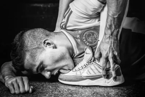 "Yearning" by Arjan Spannenburg: A museum-quality black and white fine art photograph exploring the profound emotional depth of longing and the human condition, depicting a man in an Ajax jersey licking a sneaker in a display of radical desire and devotion.